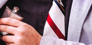 A close-up of a man’s chest wearing a black shirt and a striped tie, hiding a small flask under his grey suit jacket.