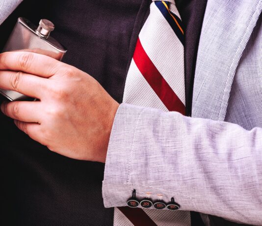 A close-up of a man’s chest wearing a black shirt and a striped tie, hiding a small flask under his grey suit jacket.