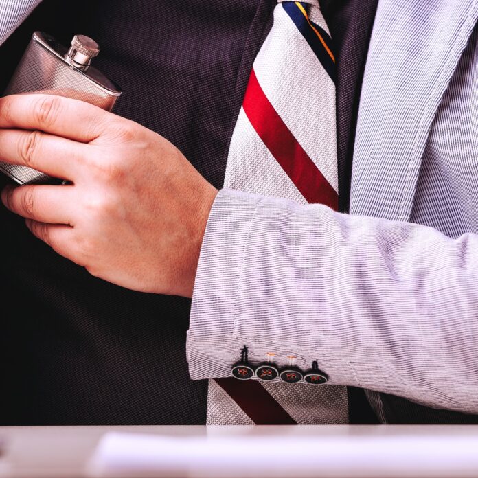 A close-up of a man’s chest wearing a black shirt and a striped tie, hiding a small flask under his grey suit jacket. A close-up of a man’s chest wearing a black shirt and a striped tie, hiding a small flask under his grey suit jacket.