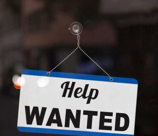 A small blue-and-white sign suction-cupped to a glass surface. The sign has text reading "Help Wanted" on it.