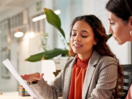 Two businesswomen sitting at the same desk. They are looking at a piece of paper in one of their hands.