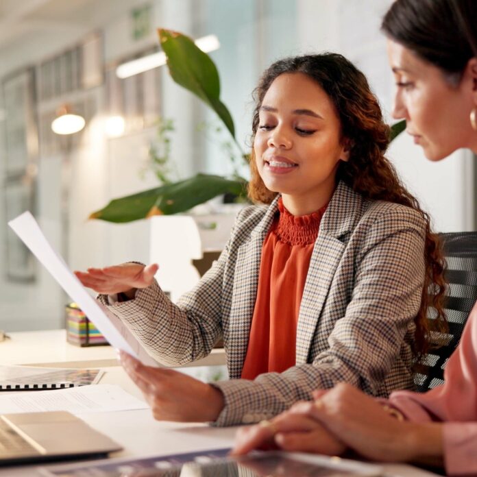 Two businesswomen sitting at the same desk. They are looking at a piece of paper in one of their hands. Two businesswomen sitting at the same desk. They are looking at a piece of paper in one of their hands.