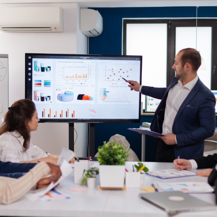 A man wearing a blue suit jacket, presenting on a large monitor in front of a room with people sitting on a desk. A man wearing a blue suit jacket, presenting on a large monitor in front of a room with people sitting on a desk.