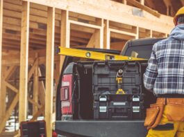 A man wearing yellow pants and a tool belt, standing in the back of a pickup truck parked on a construction site.