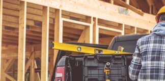 A man wearing yellow pants and a tool belt, standing in the back of a pickup truck parked on a construction site.