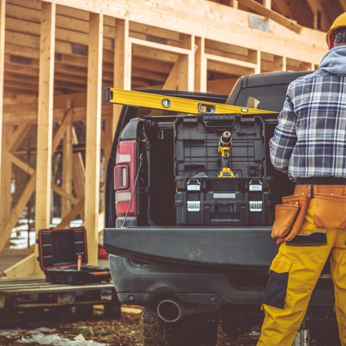 A man wearing yellow pants and a tool belt, standing in the back of a pickup truck parked on a construction site. A man wearing yellow pants and a tool belt, standing in the back of a pickup truck parked on a construction site.