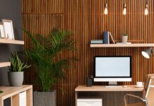 A home office features a wooden slat accent wall, a desk with a computer, a tan office chair, and several potted plants.