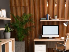 A home office features a wooden slat accent wall, a desk with a computer, a tan office chair, and several potted plants.