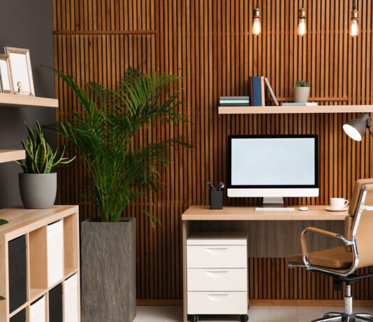 A home office features a wooden slat accent wall, a desk with a computer, a tan office chair, and several potted plants.