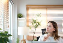 A woman appears to be in deep thought as she looks out the window of her office. Her laptop sits in front of her.