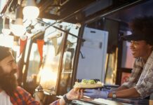 What To Budget Before Buying a Mobile Shop A woman in gloves handing a sandwich to a bearded man at a food truck window in warm outdoor light as they smile.