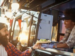 A woman in gloves handing a sandwich to a bearded man at a food truck window in warm outdoor light as they smile.