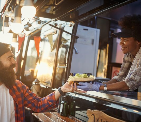 A woman in gloves handing a sandwich to a bearded man at a food truck window in warm outdoor light as they smile.