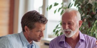 A father explains his business to his younger son at a home office table with papers, a laptops, mugs, and a plant.