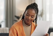 A remote business owner reviews paperwork while taking notes and working on a laptop from her bright home office.