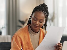 A remote business owner reviews paperwork while taking notes and working on a laptop from her bright home office.