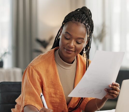 A remote business owner reviews paperwork while taking notes and working on a laptop from her bright home office.