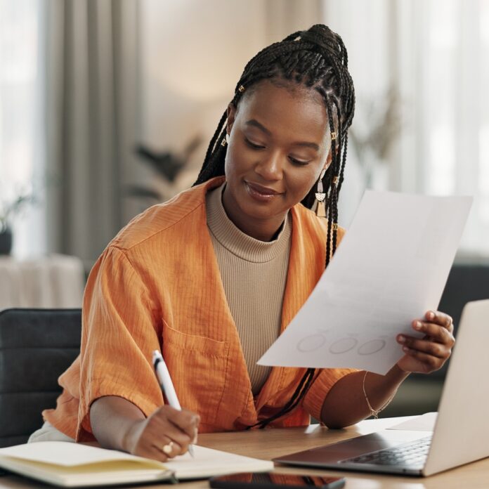 A remote business owner reviews paperwork while taking notes and working on a laptop from her bright home office.