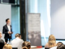 A group of business professionals all seated in front of a speaker who is standing on a stage in front of them.