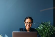 A smiling woman works on a laptop at a wooden desk with pieces of paper and green plants in a blue office.