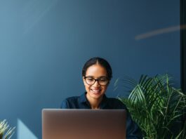 A smiling woman works on a laptop at a wooden desk with pieces of paper and green plants in a blue office.