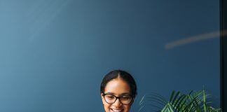 A smiling woman works on a laptop at a wooden desk with pieces of paper and green plants in a blue office.