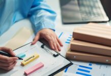 The Operational Bottlenecks Growing Businesses Miss A person in a blue striped shirt writes on a clipboard beside sticky notes, charts, folders, and a laptop.