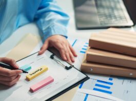 A person in a blue striped shirt writes on a clipboard beside sticky notes, charts, folders, and a laptop.