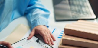 A person in a blue striped shirt writes on a clipboard beside sticky notes, charts, folders, and a laptop.