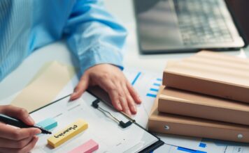 The Operational Bottlenecks Growing Businesses Miss A person in a blue striped shirt writes on a clipboard beside sticky notes, charts, folders, and a laptop.