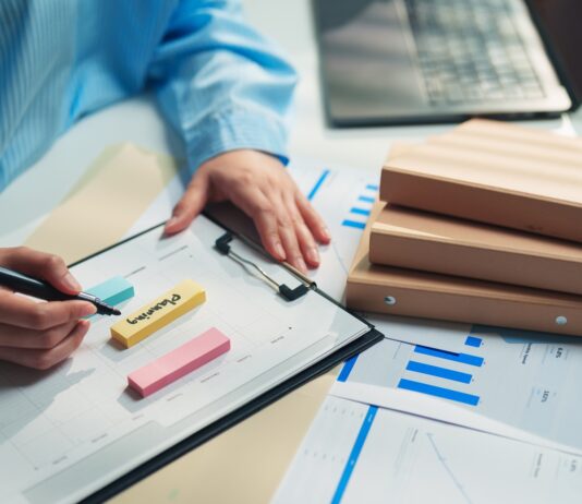 A person in a blue striped shirt writes on a clipboard beside sticky notes, charts, folders, and a laptop.