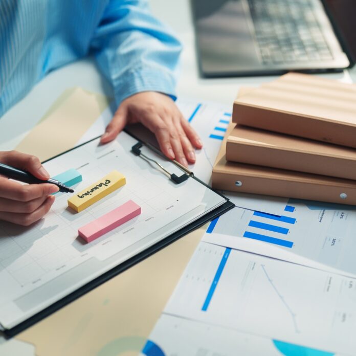 A person in a blue striped shirt writes on a clipboard beside sticky notes, charts, folders, and a laptop. A person in a blue striped shirt writes on a clipboard beside sticky notes, charts, folders, and a laptop.